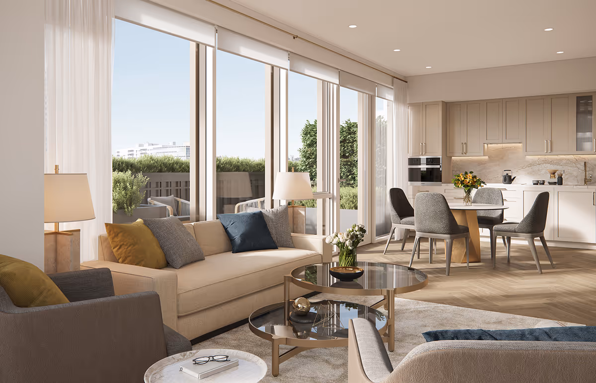 A bright and modern living room and kitchen area with large floor-to-ceiling windows letting in natural light. The living room features a beige sofa with colorful cushions, a round glass coffee table with decorative items, and a side table with a lamp. The kitchen area has light-colored cabinets, a round dining table with four chairs, and a vase with flowers on the table. Outside the windows, greenery is visible on a balcony or patio.