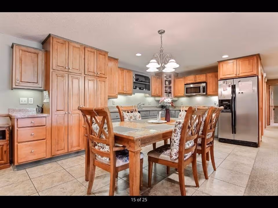 A spacious kitchen with wooden cabinets, a stainless steel refrigerator, microwave, and a wooden dining table with six chairs featuring floral cushions. A chandelier with five lights hangs above the table, and the floor is tiled.