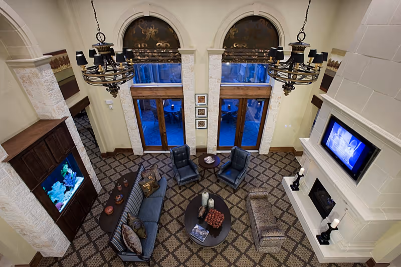 A spacious and elegant living room with patterned carpet, a large sofa, two armchairs, a bench, and a coffee table with decorative items. The room features tall arched windows with wooden frames, two ornate chandeliers, a built-in aquarium, and a wall-mounted television above a fireplace.