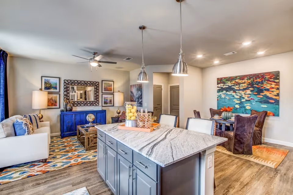 A bright and modern open-concept living space featuring a kitchen island with a marble countertop and two pendant lights above. The living room area has a white sofa, a blue sideboard with a decorative mirror and framed artwork on the wall, and a colorful patterned rug. The dining area includes a round table with four chairs and a large colorful painting on the wall. The flooring is wood, and the walls are painted light gray.