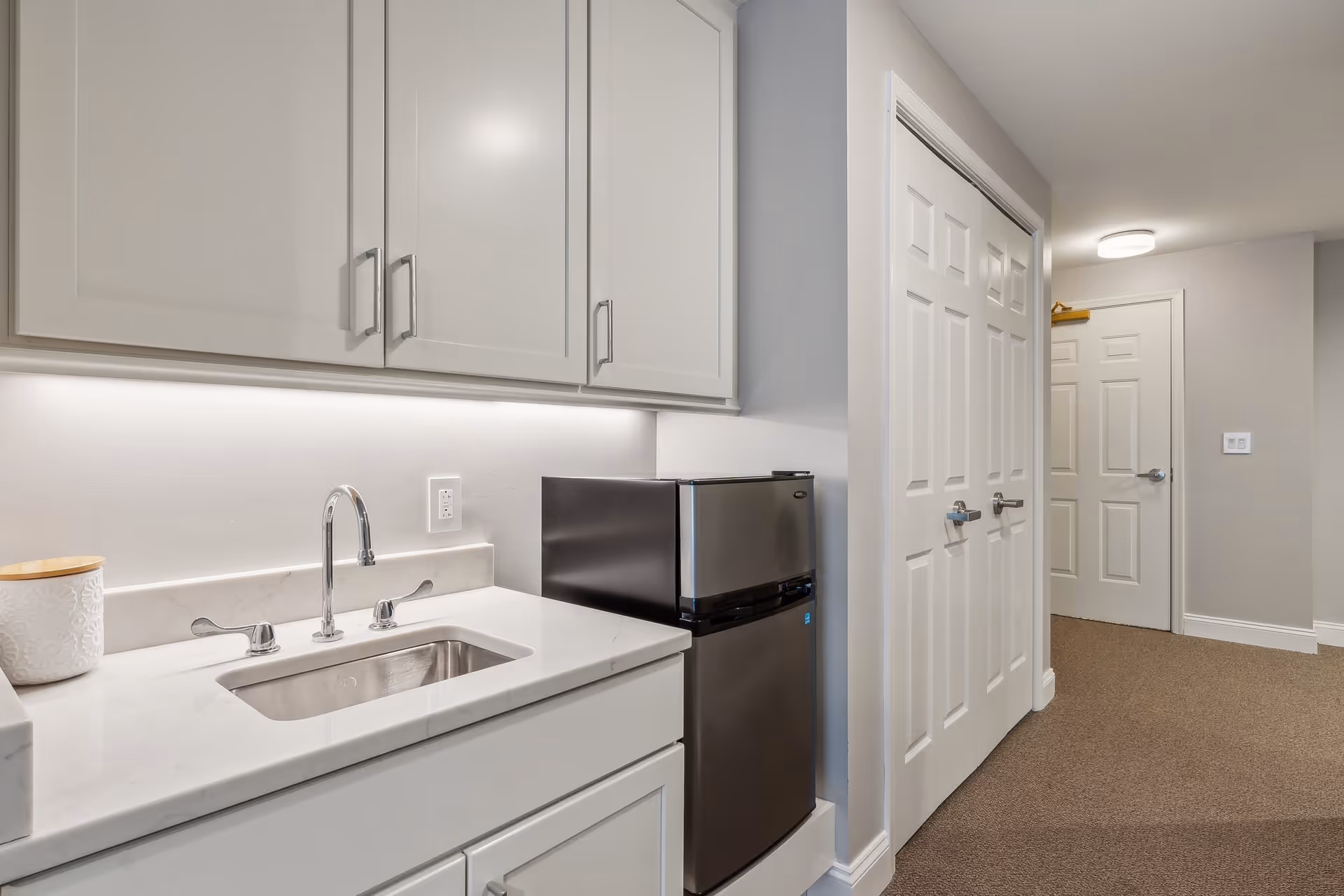 A small kitchenette area with white cabinets, a stainless steel sink with a chrome faucet, a small stainless steel refrigerator, and a white countertop. The walls are painted light gray, and there is a hallway with white doors and beige carpet visible in the background.