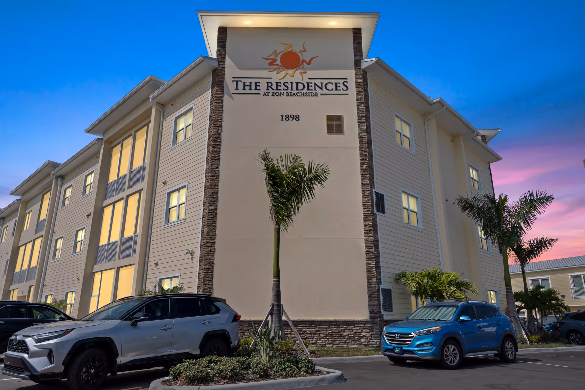 Exterior front of a three-story building labeled "The Residences at Zon Beachside" with parked cars and palm trees at dusk.