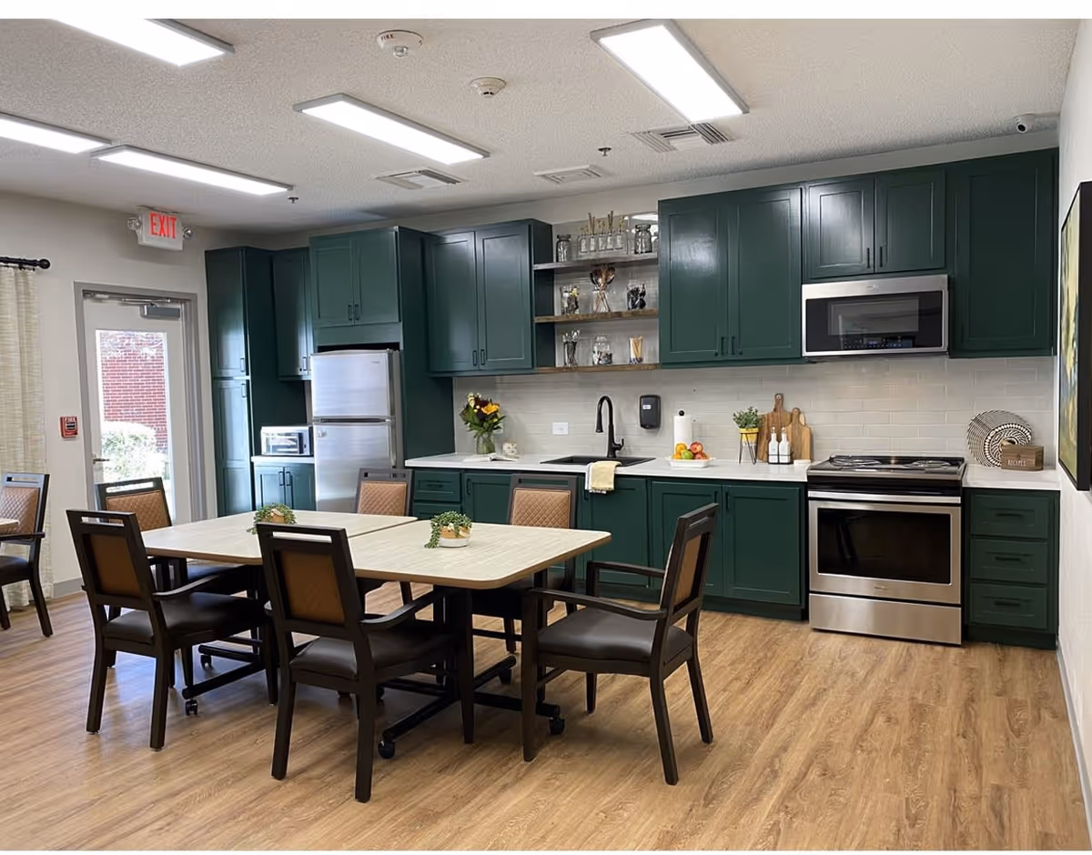 A modern kitchen and dining area with green cabinets, stainless steel appliances including a refrigerator, microwave, and oven. There is a rectangular dining table with six chairs, small potted plants on the table, and wooden flooring. The room is well-lit with ceiling lights and has a door with a window on the left side.