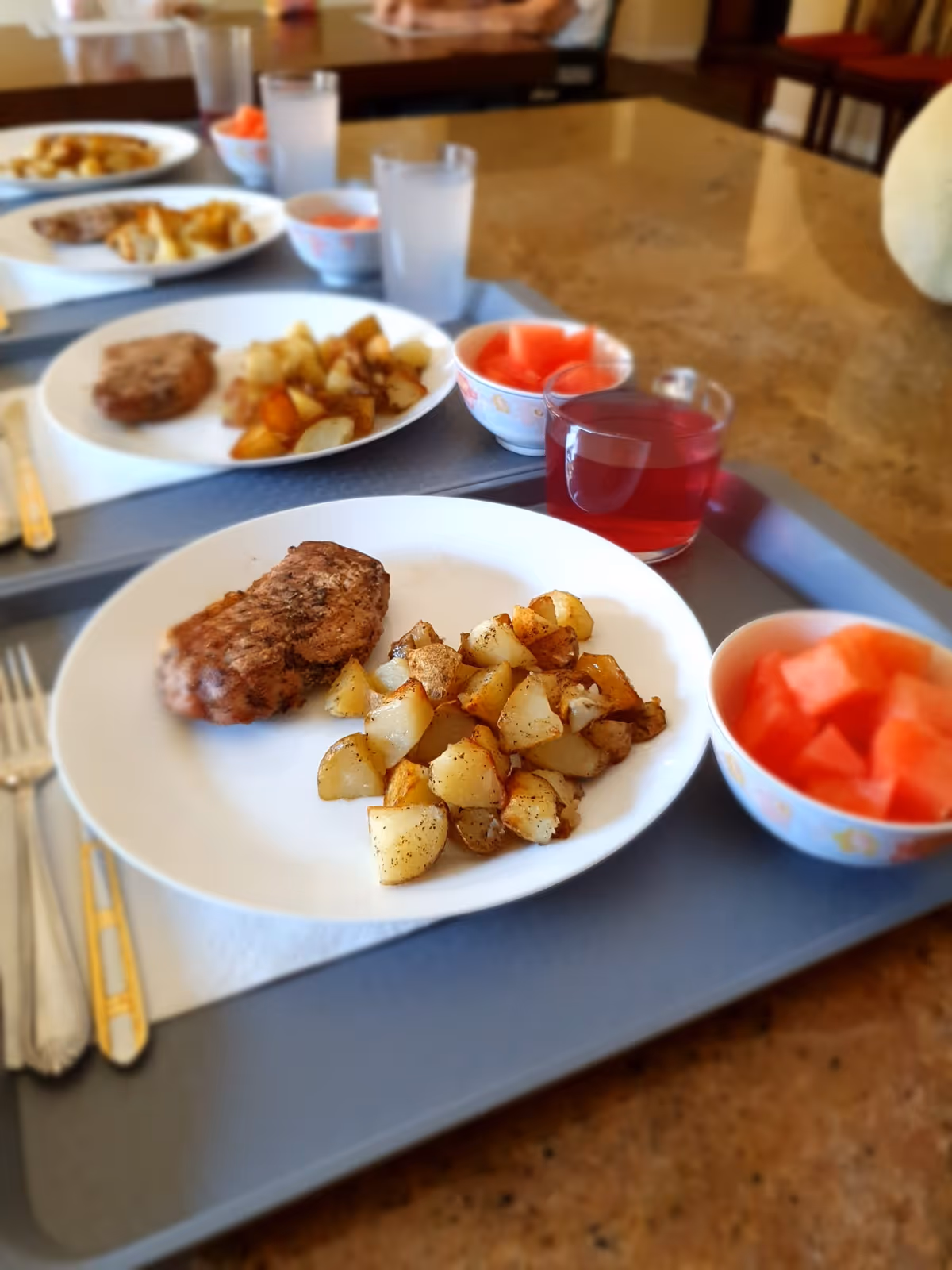 A meal on trays with a plate of meat and roasted potatoes, bowls of watermelon, and cups on a dining table.