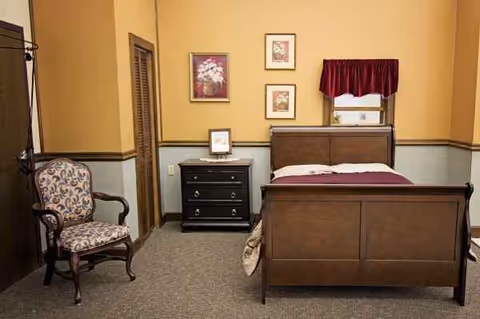 A cozy patient bedroom with wooden bed, dresser, chair, and framed artwork.