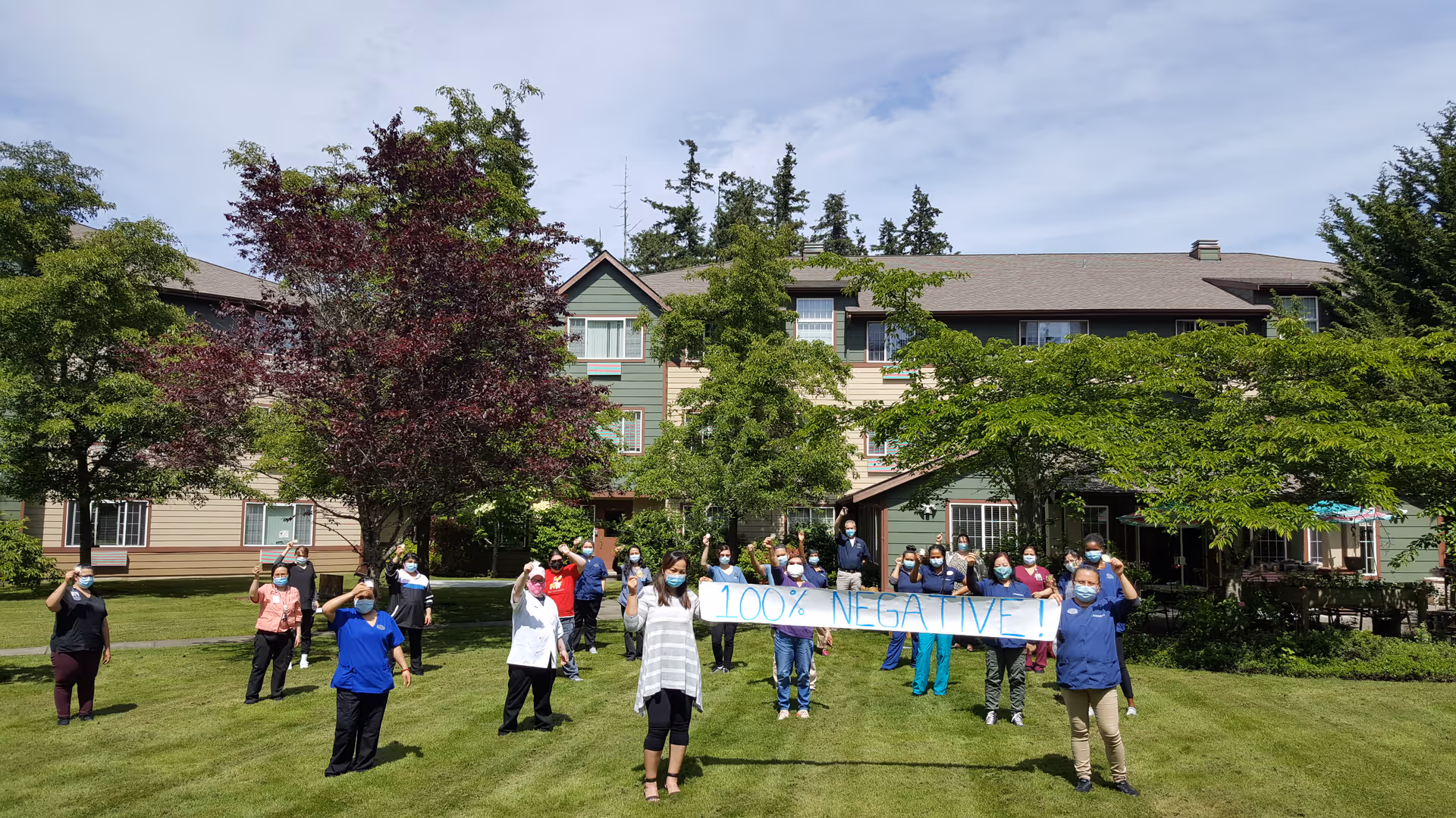 Group of masked staff standing on a lawn in front of a multi-story senior living building holding a banner that reads "100% NEGATIVE".