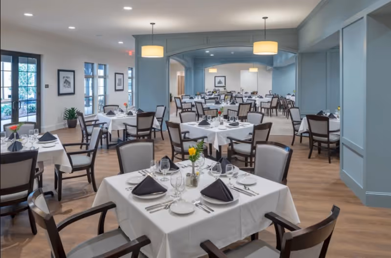 A spacious dining room with multiple tables covered in white tablecloths, each set with plates, silverware, glasses, and black folded napkins. The room features wooden chairs with cushioned seats, light blue walls with paneling, pendant lights hanging from the ceiling, and large windows letting in natural light. There are framed pictures on the walls and small flower arrangements on each table.