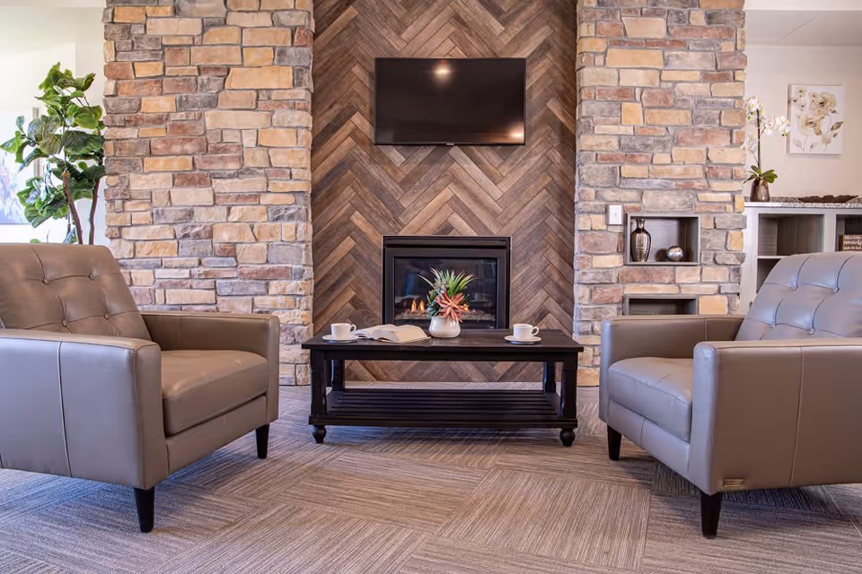 A cozy living room area with two gray leather armchairs facing a black coffee table. The table holds a small plant, an open book, and two white coffee cups. Behind the table is a fireplace with a wooden herringbone pattern wall and a mounted flat-screen TV above it. The room features stone pillars on either side of the fireplace and decorative items on shelves to the right.