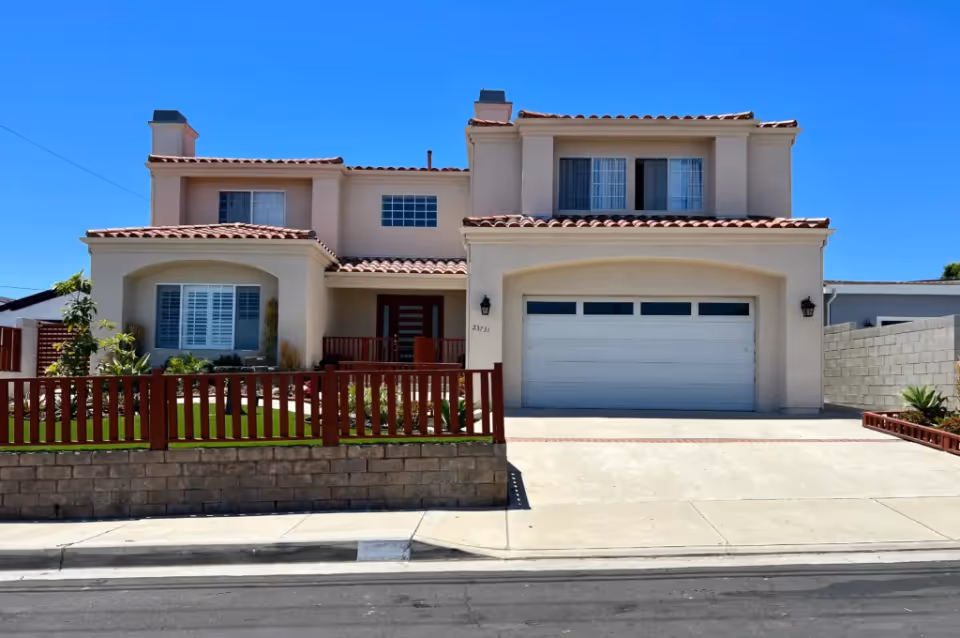 Two-story stucco house with a tiled roof, attached two-car garage, red wooden fence and a small front yard under a clear blue sky.