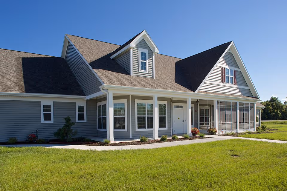 Exterior view of a single-story residential building with gray siding, white trim, multiple windows, a front porch with white columns, and a well-maintained green lawn under a clear blue sky.