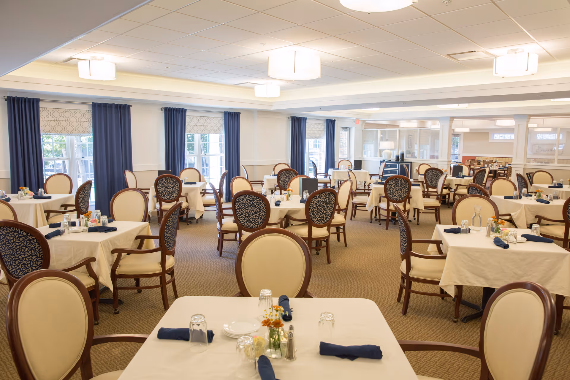 A spacious dining room with multiple tables covered in white tablecloths, each set with plates, glasses, napkins, and small flower arrangements. The room has large windows with blue curtains and beige patterned carpet, and elegant wooden chairs with cream and dark patterned upholstery.