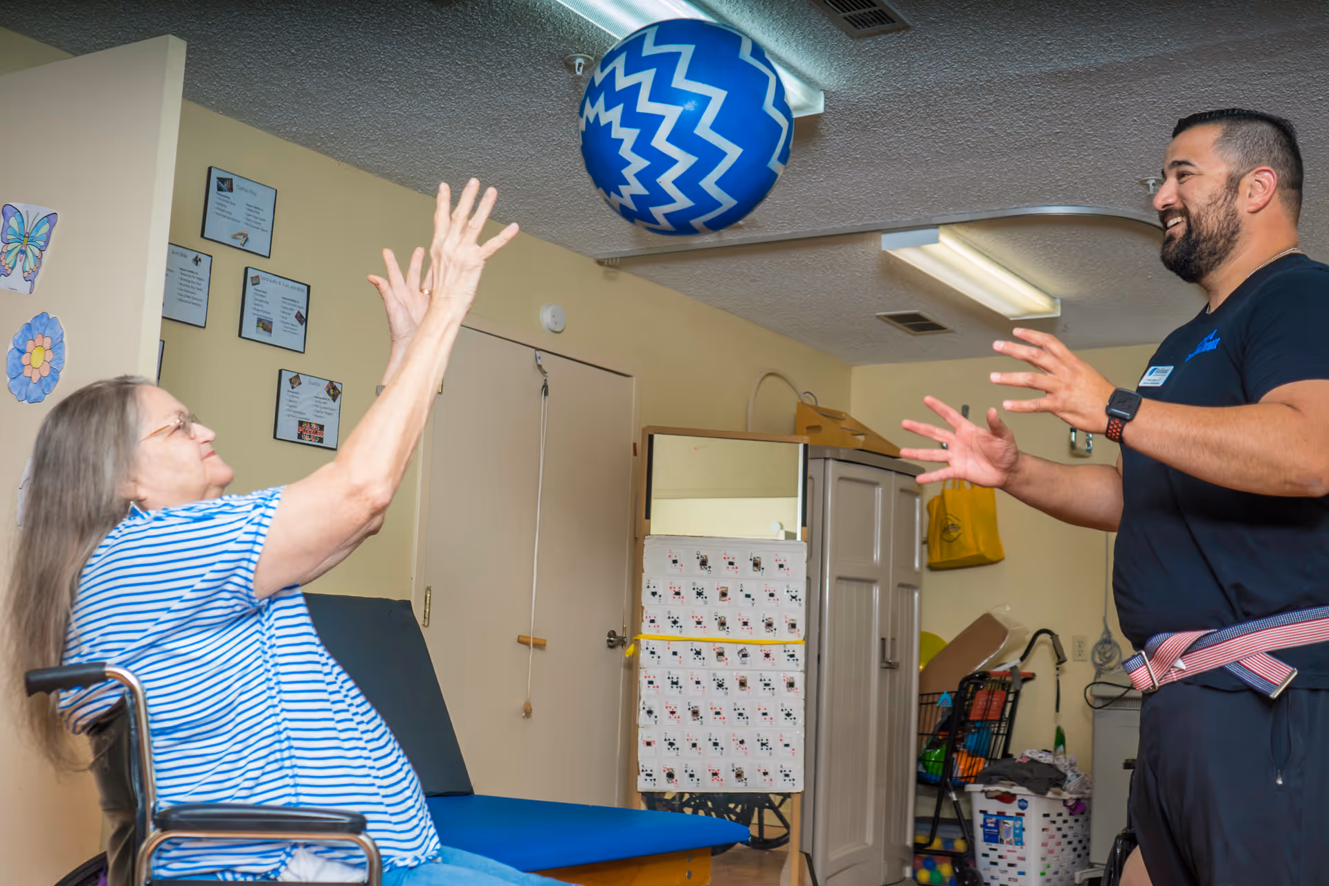 An elderly woman in a wheelchair playing with a large blue and white patterned ball with a smiling man in a black shirt in a room that appears to be a therapy or activity area.