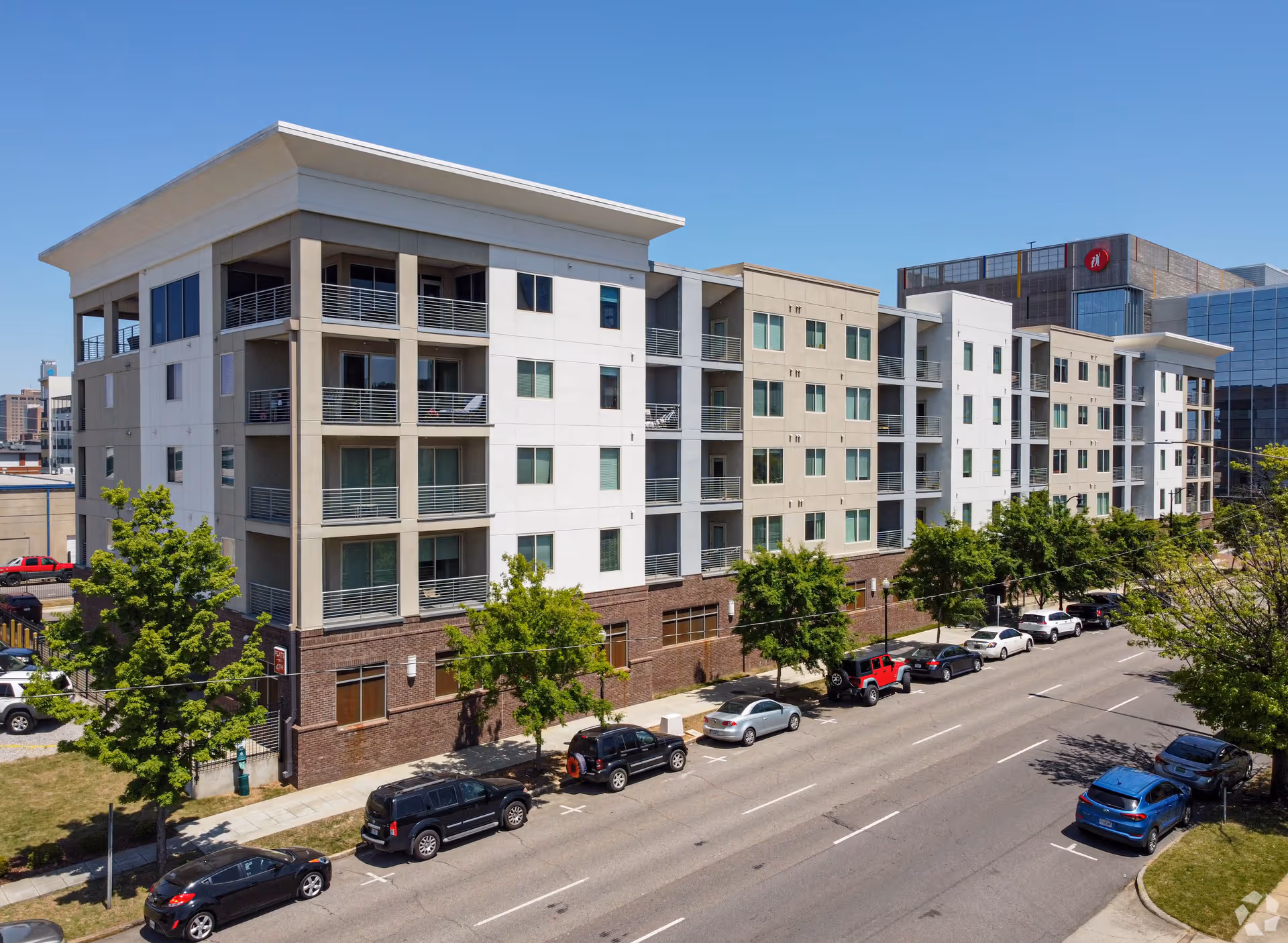 Exterior view of a modern multi-story residential building with balconies, situated along a street with parked cars and trees under a clear blue sky.
