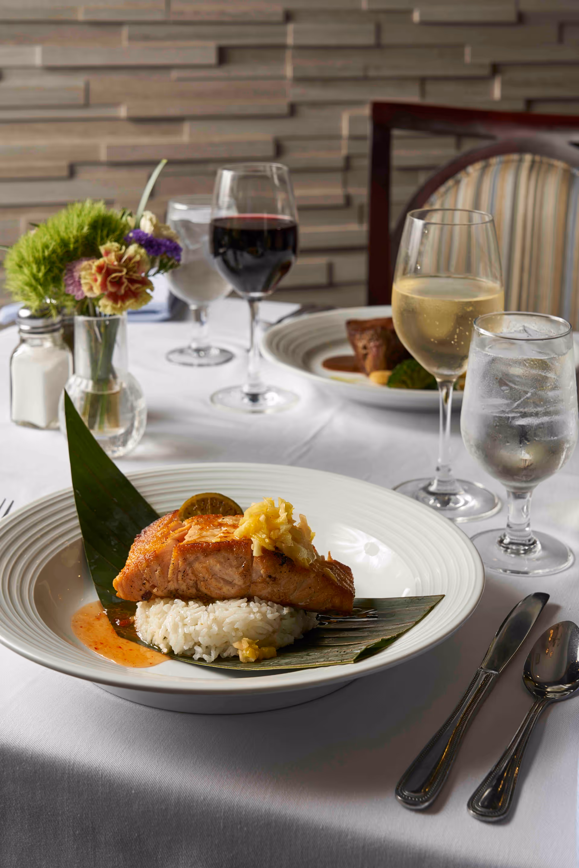 A plated salmon fillet on rice atop a leaf on a white tablecloth with wine and water glasses and place settings in the background.