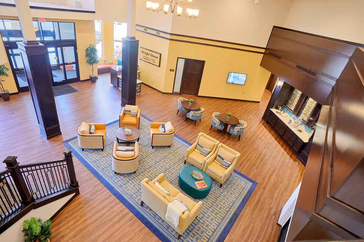 A spacious and well-lit senior living facility common area with wooden floors, comfortable seating including armchairs and sofas arranged around coffee tables on a large patterned rug. There are tall dark wooden columns, a staircase with a wooden railing, and a wall-mounted TV. The walls are painted yellow and white, and there is a sign that reads 'Wright House Memory Care'.