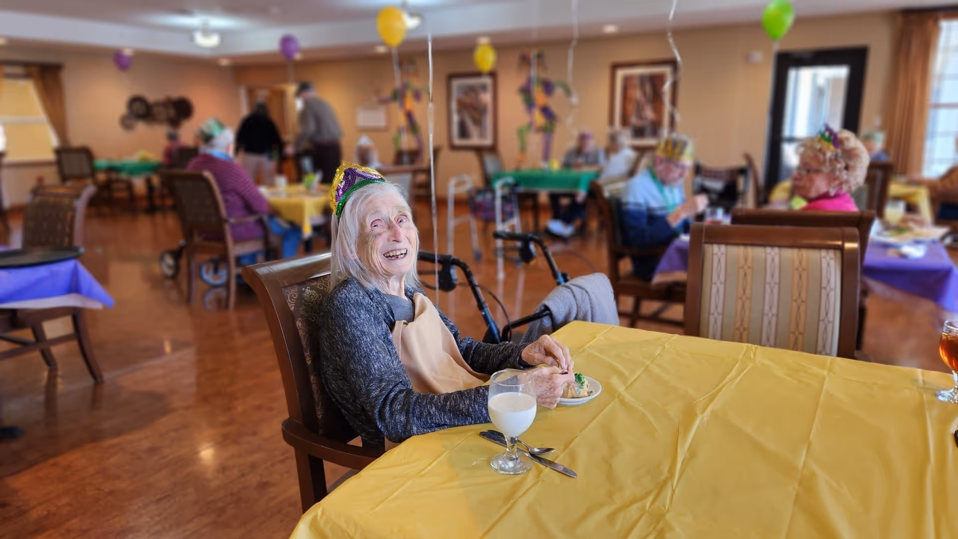 An elderly woman wearing a party hat sits at a table with a yellow tablecloth, smiling and eating a dessert with a glass of milk in front of her. Other elderly people are seated at tables decorated with colorful balloons in the background, suggesting a festive gathering in a dining area.