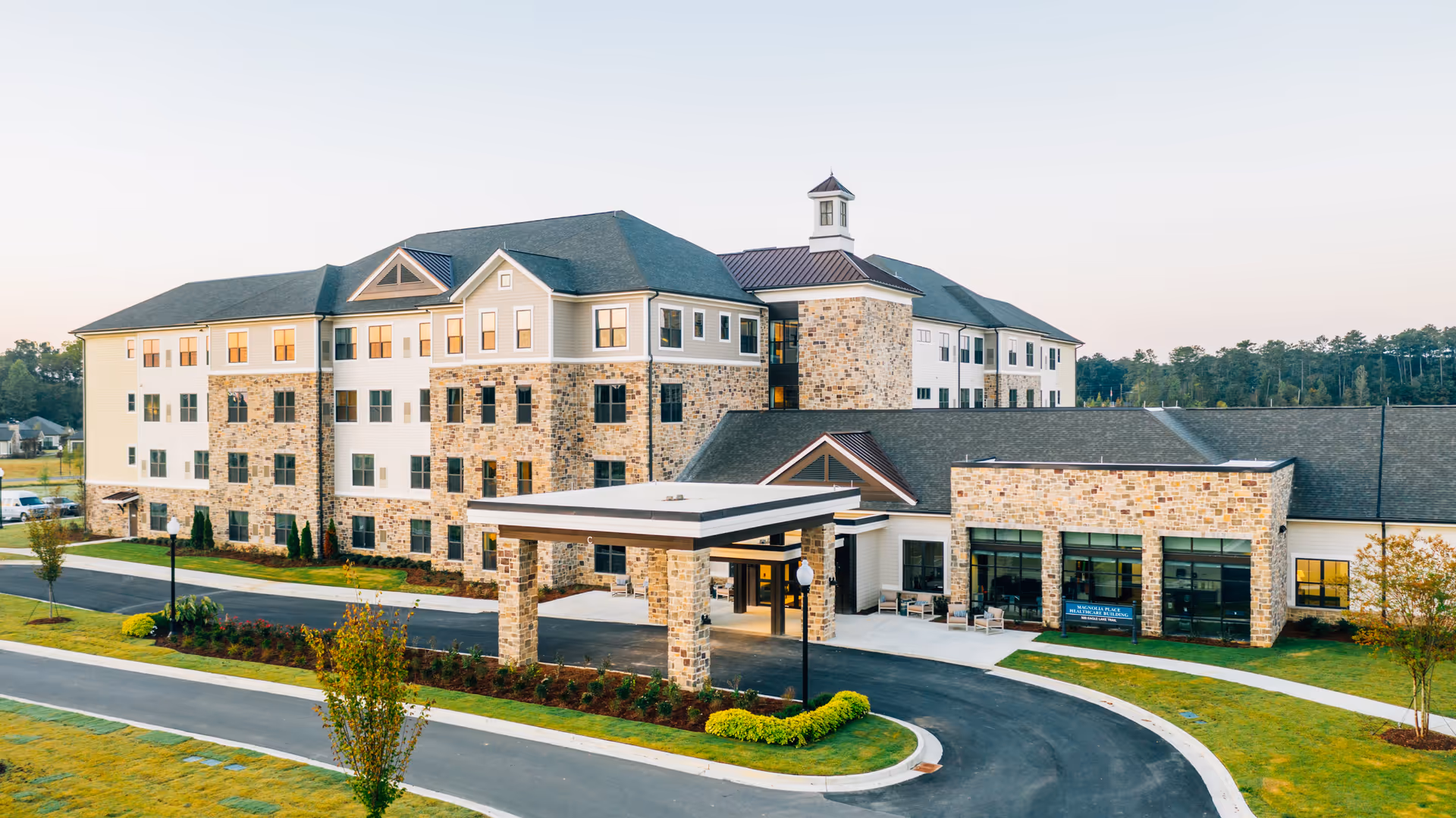 Exterior view of a large, multi-story senior living facility with stone and siding facade, a covered entrance, landscaped grounds, and a circular driveway.