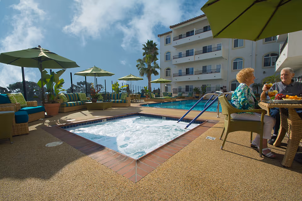 Outdoor patio area at San Clemente Villas featuring a bubbling hot tub in the foreground, a swimming pool behind it, several green umbrellas providing shade over wicker seating with colorful cushions, and two elderly people sitting at a table enjoying drinks and snacks under an umbrella. A multi-story building with balconies is visible in the background under a partly cloudy sky.