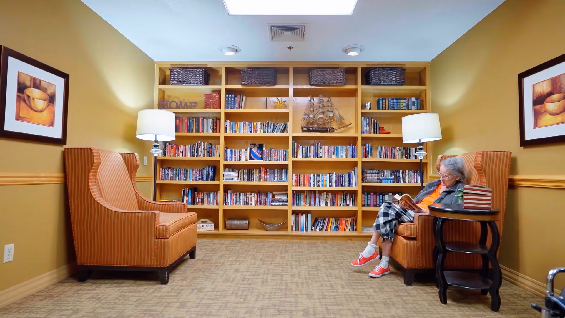 Cozy reading room with built-in bookshelves, two upholstered armchairs and lamps, and an elderly woman reading a book.