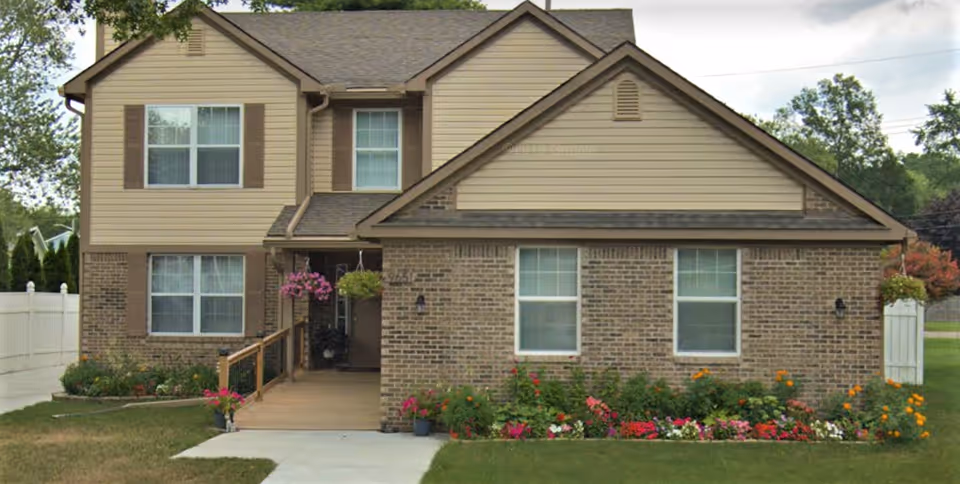 Front exterior view of a two-story residential building with beige siding and brick facade, featuring multiple windows with brown shutters, a small porch with hanging flower baskets, a wooden ramp, and a well-maintained garden with colorful flowers in front.