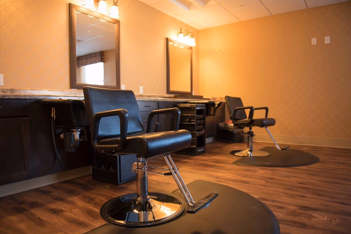 Interior view of a senior living facility's hair salon area with two black salon chairs on circular mats, large mirrors mounted on a beige patterned wall, and dark wood cabinetry with granite countertops beneath the mirrors.
