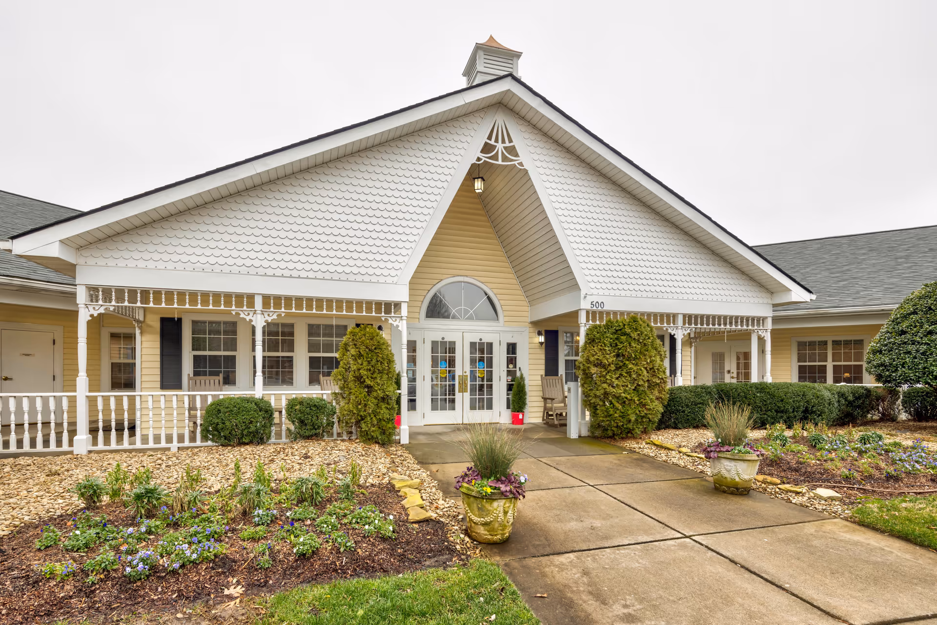 Front exterior view of a single-story building with a peaked roof and white siding. The entrance features double glass doors with sidelights, flanked by bushes and potted plants. There is a covered porch area with white railings and decorative trim. The building number 500 is visible near the entrance.