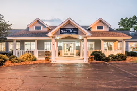Front entrance of a single-story senior living building with a covered porch, white columns, and a sign that reads 'Welcome to Seneca House'.