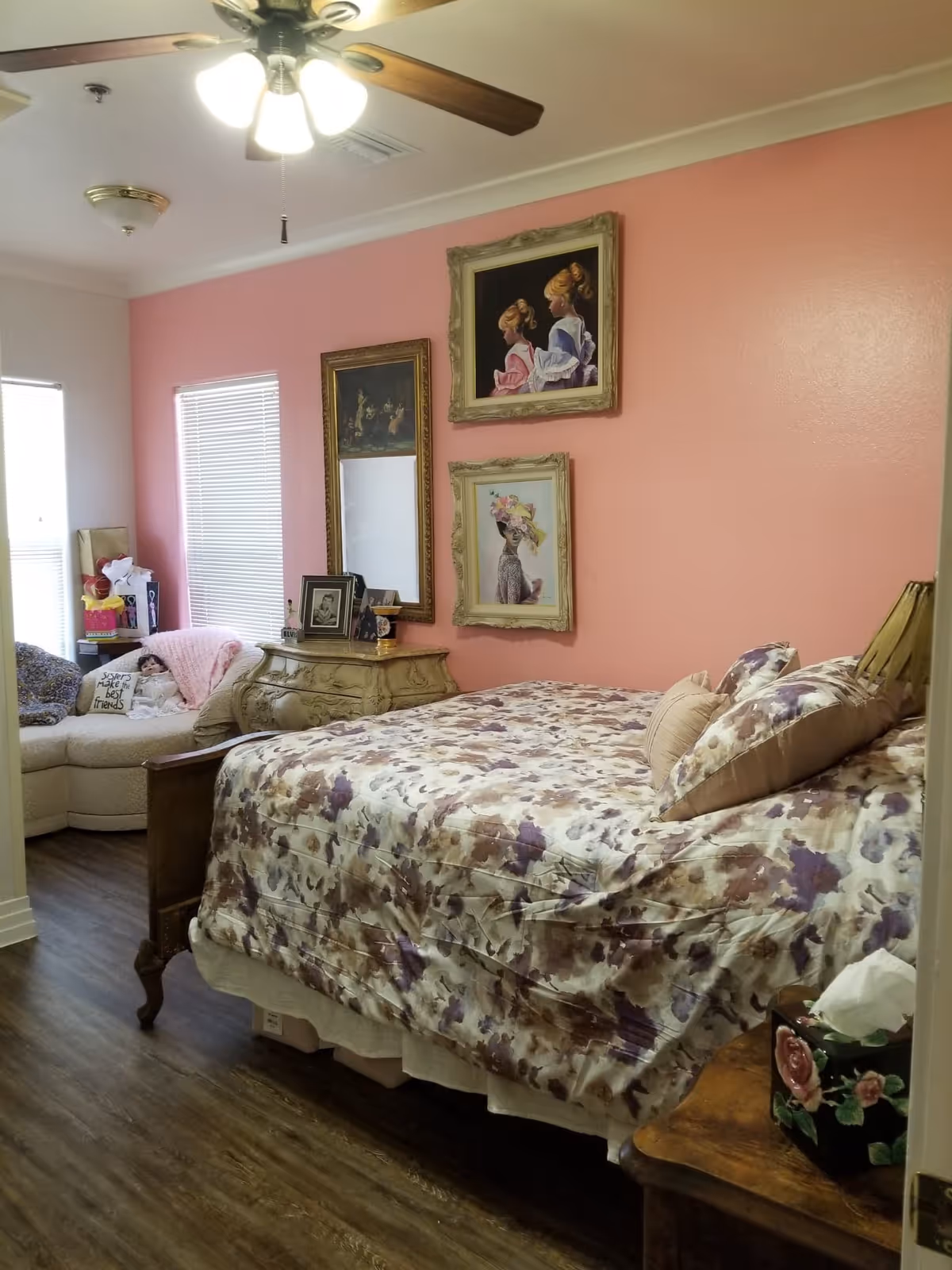 A bedroom with pink walls featuring three framed paintings, a floral bedspread on a wooden bed, a wooden nightstand with a floral tissue box, a decorative dresser with framed photos, and a cozy corner seating area with a doll and a pillow that says 'Sisters make the best friends'.