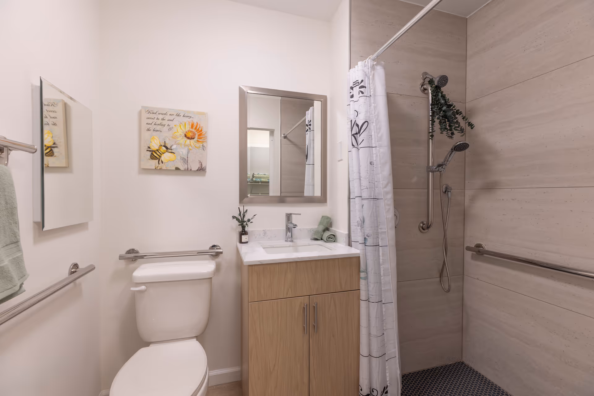 A clean and modern bathroom featuring a white toilet, a wooden vanity with a white countertop and a rectangular mirror above it. There is a shower area with beige tiles, a handheld showerhead, and a white shower curtain with black patterns. The bathroom also has safety grab bars and a small decorative plant on the vanity.