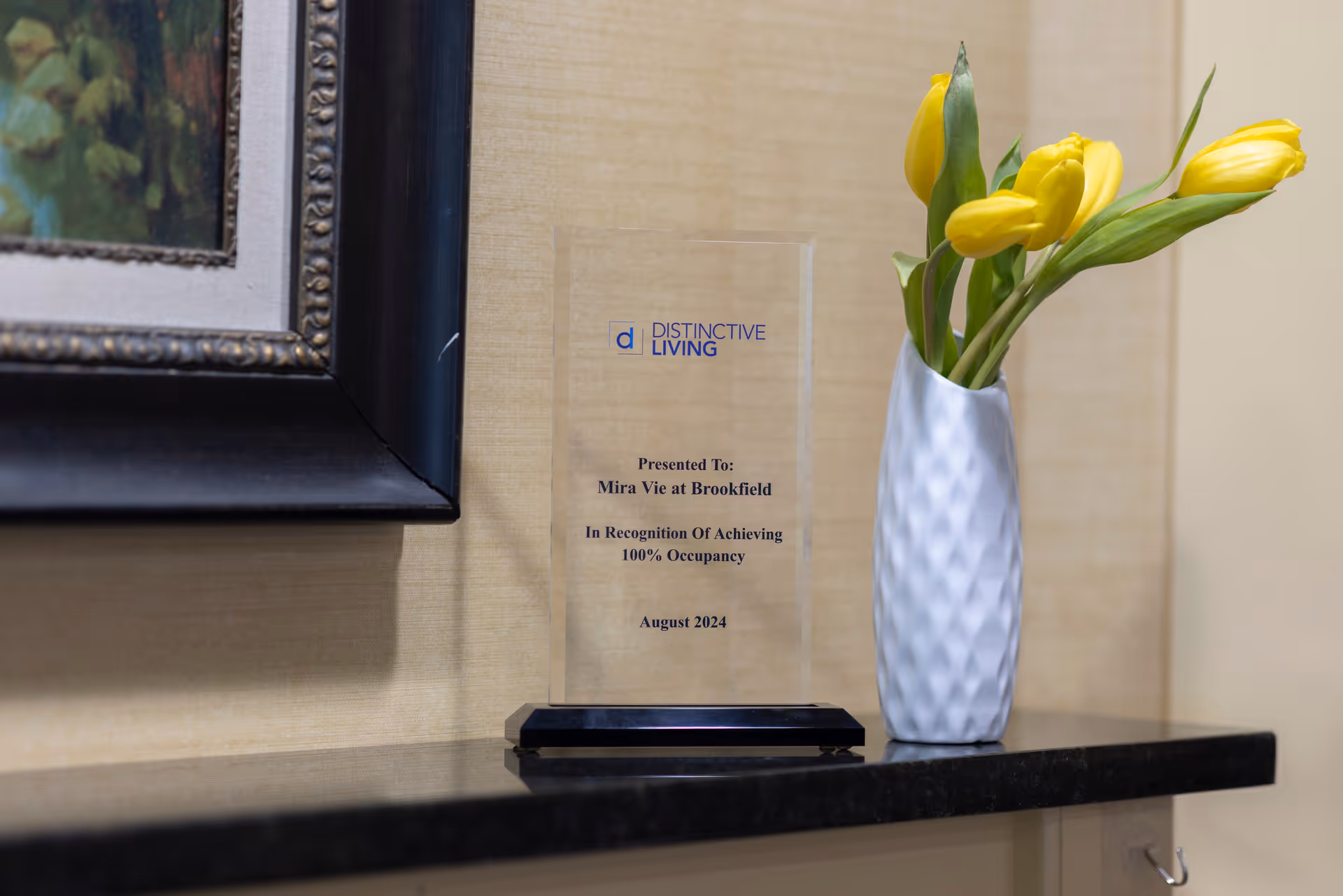 Clear acrylic award plaque on a black countertop next to a white vase of yellow tulips and part of a framed painting.