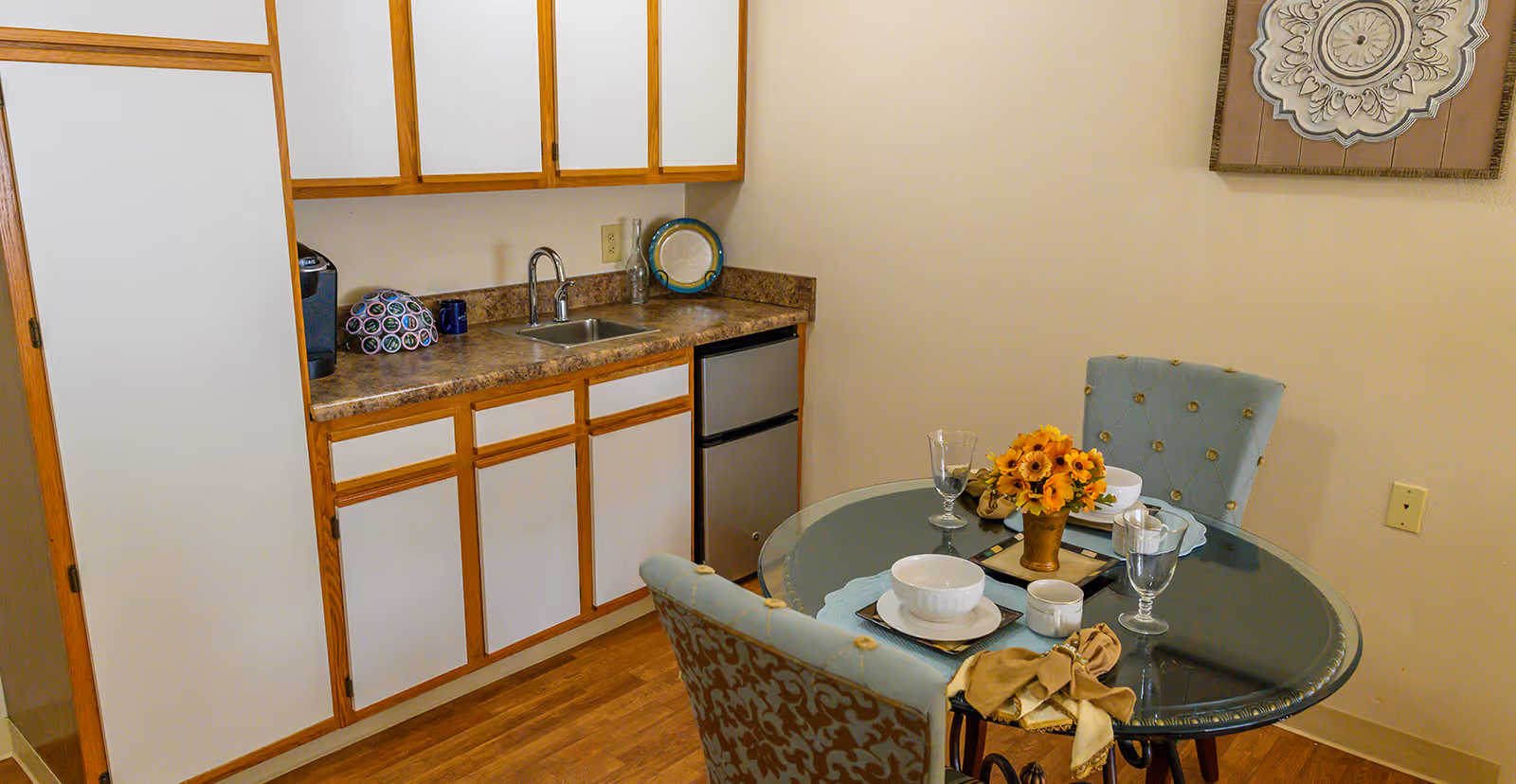 A small kitchen area with white cabinets and wooden trim, a countertop with a sink, a coffee maker, and a mini refrigerator. Next to the kitchen is a round glass dining table set for two with blue upholstered chairs, white bowls, glasses, and a small vase with yellow flowers. A decorative wall art piece is visible on the beige wall.