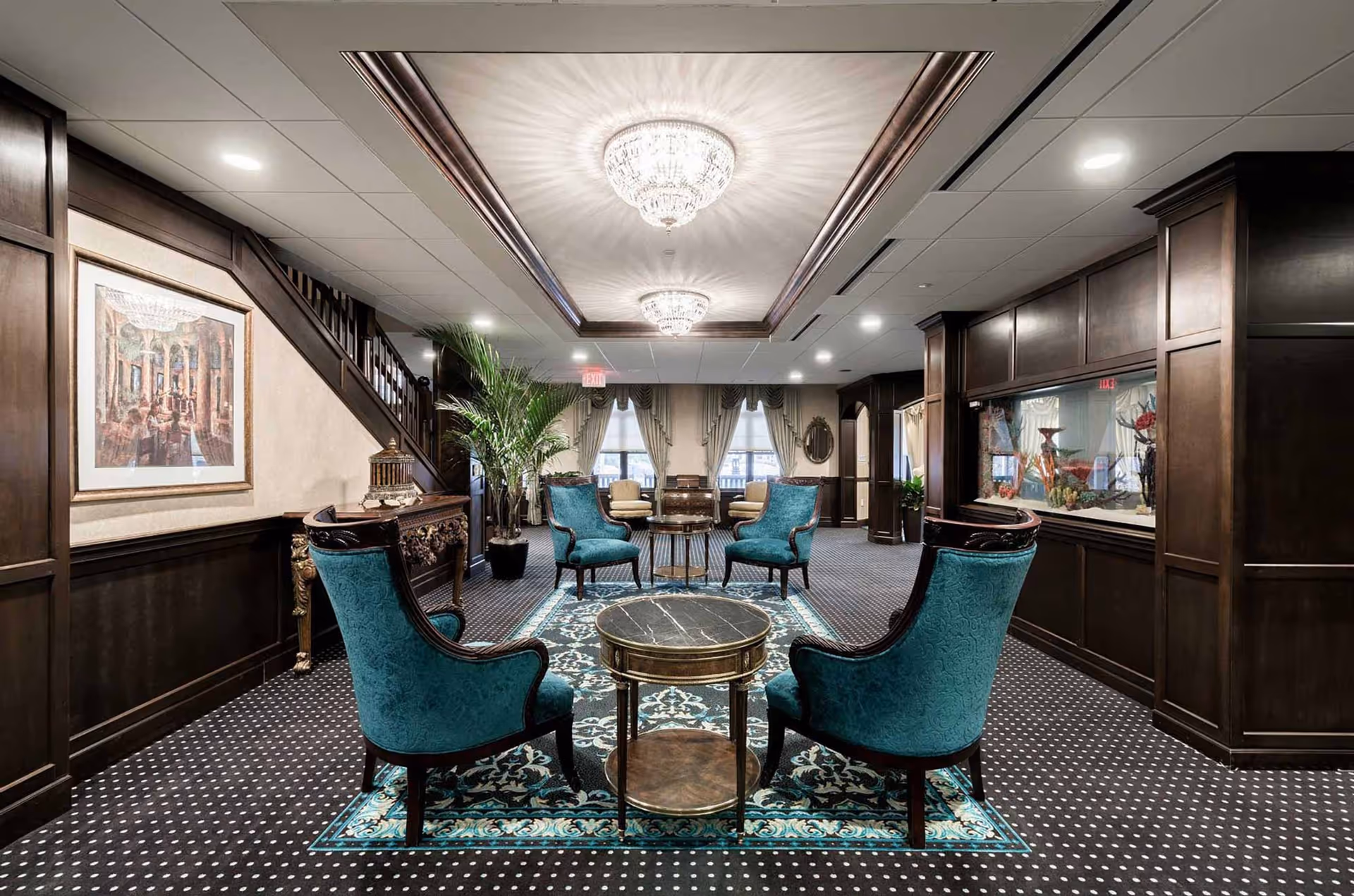 An elegant interior lounge area with dark wood paneling and trim, featuring four teal upholstered armchairs arranged around a round marble-top coffee table on a patterned rug. The ceiling has recessed lighting and two ornate chandeliers. There are large windows with draped curtains in the background and decorative plants and artwork on the walls.