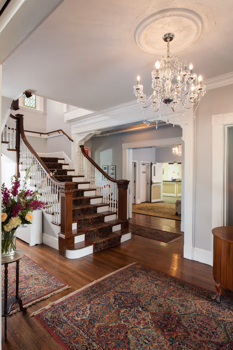 Interior view of a nursing center featuring a wooden staircase with a carpet runner, ornate chandelier hanging from the ceiling, floral arrangements on a small table, and multiple patterned rugs on the hardwood floor. The space has light-colored walls and an open doorway leading to another room.
