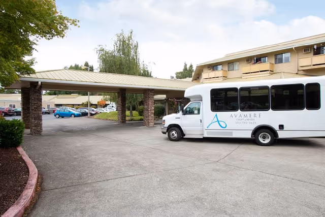Exterior view of Avamere Court at Keizer showing a covered driveway area with stone pillars and a white shuttle bus parked nearby. The building has multiple windows and balconies, and there are several cars parked in the background under a cloudy sky.
