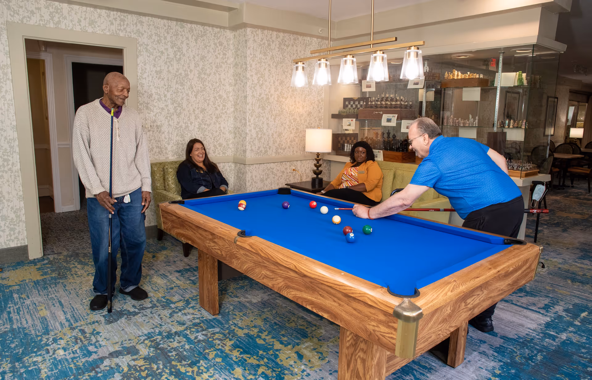 A group of four people in a recreational room at Oak Park Arms. Two women are seated on a green couch, one man is standing holding a pool cue, and another man is leaning over a pool table with a blue felt, aiming to take a shot. The room has patterned wallpaper, a modern light fixture above the pool table, and a display case with chess sets in the background.