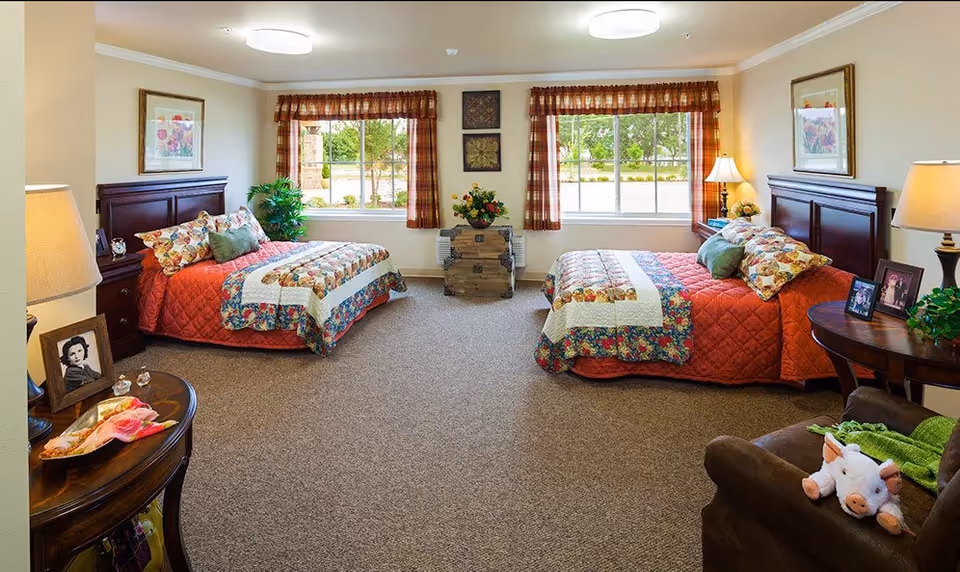A bright and cozy bedroom with two beds, each with a red quilt and floral pillows. The room has two large windows with plaid curtains, a small chest with a flower arrangement between the windows, and framed artwork on the walls. There are bedside tables with lamps and framed photos, a comfortable armchair with a stuffed pig toy, and a carpeted floor.