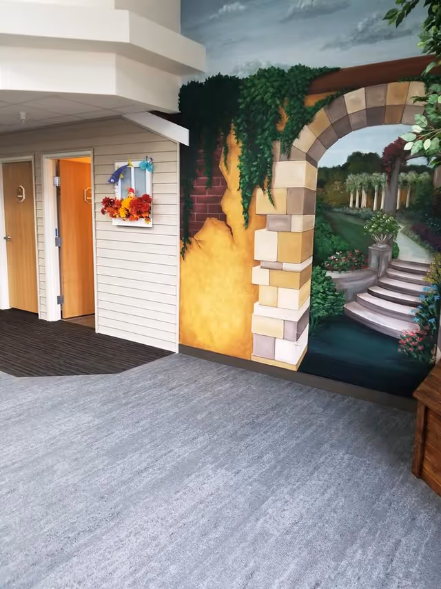 Interior hallway of a senior living facility with two wooden doors on the left, a decorative wall-mounted frame with autumn leaves, and a large mural on the right depicting a stone archway leading to a garden path with stairs and greenery.