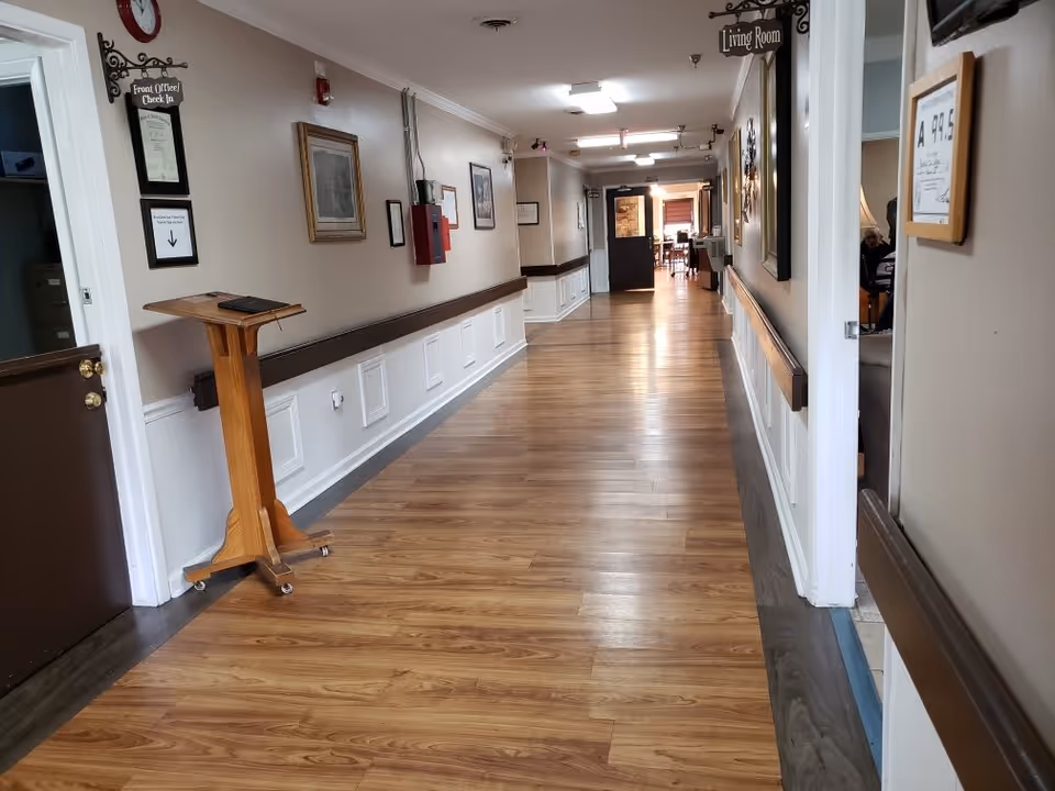 Long wood-floored interior corridor with handrails, framed artwork, and a hanging "Living Room" sign leading to a common area.