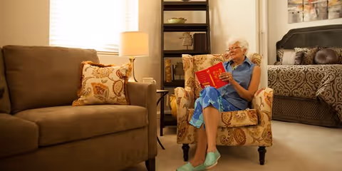 An elderly woman with short white hair sits comfortably in a patterned armchair reading a red book. She is wearing a blue dress and green slippers. The room is warmly lit with a beige sofa, a floor lamp, a tall black shelving unit, and a bed with decorative pillows in the background.