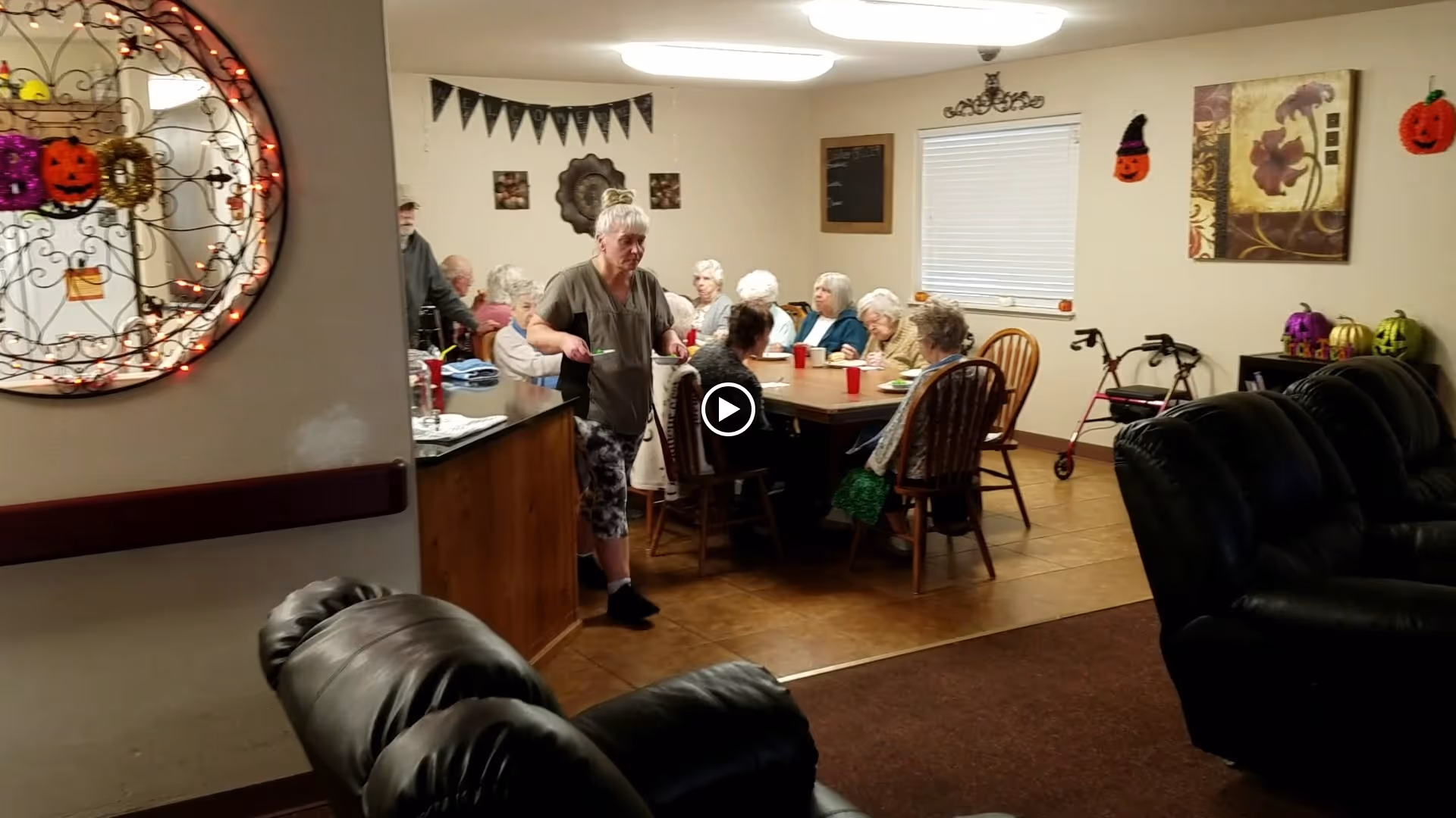 A group of elderly people sitting around a dining table in a senior care facility, with a caregiver standing nearby. The room is decorated with Halloween-themed items including pumpkin decorations on the walls and a mirror with orange lights. There are leather recliners in the foreground and a walker near the wall.