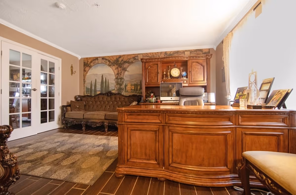 A warmly furnished living room/office with a large wooden executive desk and chair, ornate sofa, wall mural and French doors.