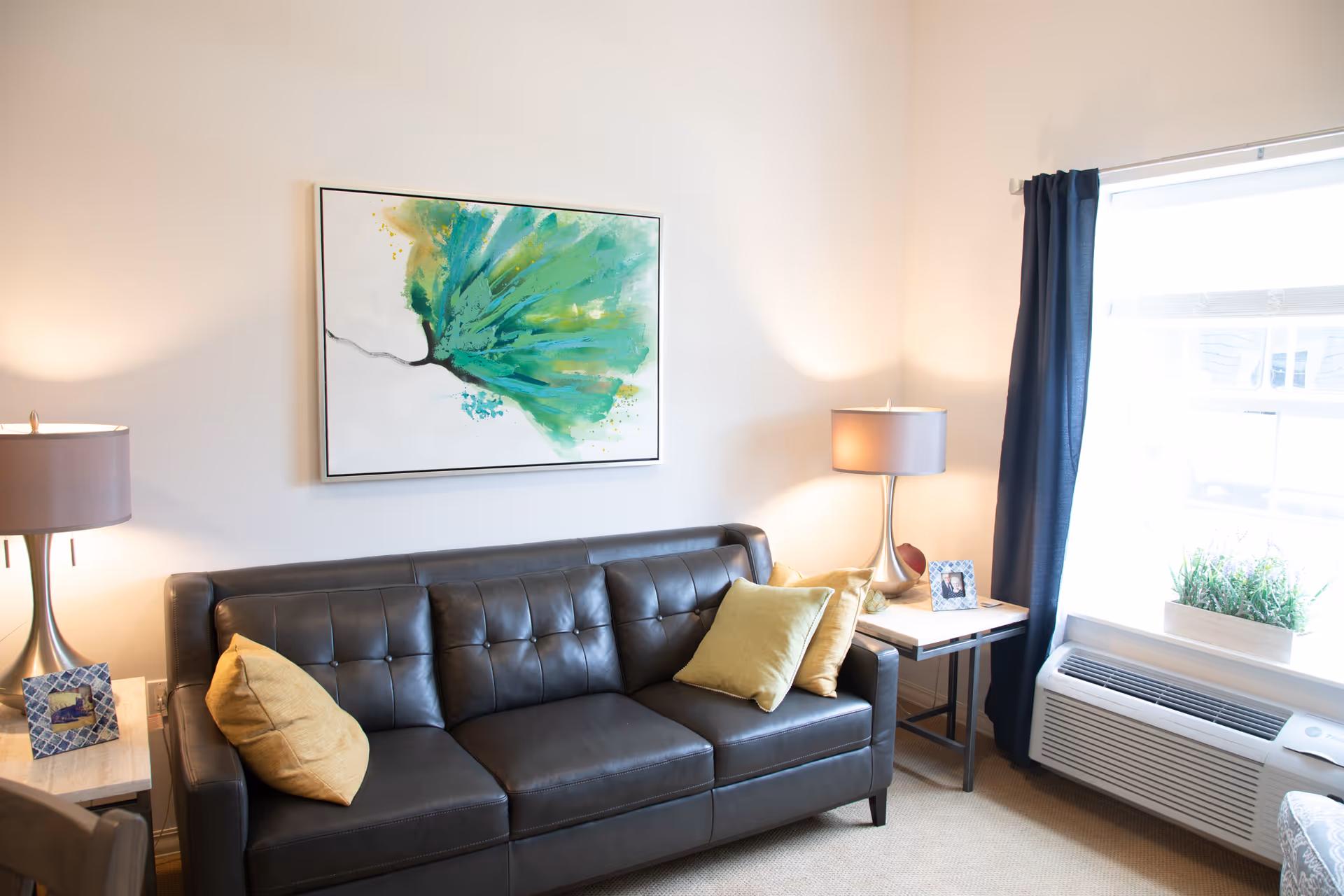 A cozy living room with a dark leather sofa adorned with yellow and green pillows. On either side of the sofa are small side tables with modern lamps and framed photos. Above the sofa hangs a large abstract painting with green and blue hues. A window with dark curtains lets in natural light, and below it is a white air conditioning unit with a planter box containing green plants.