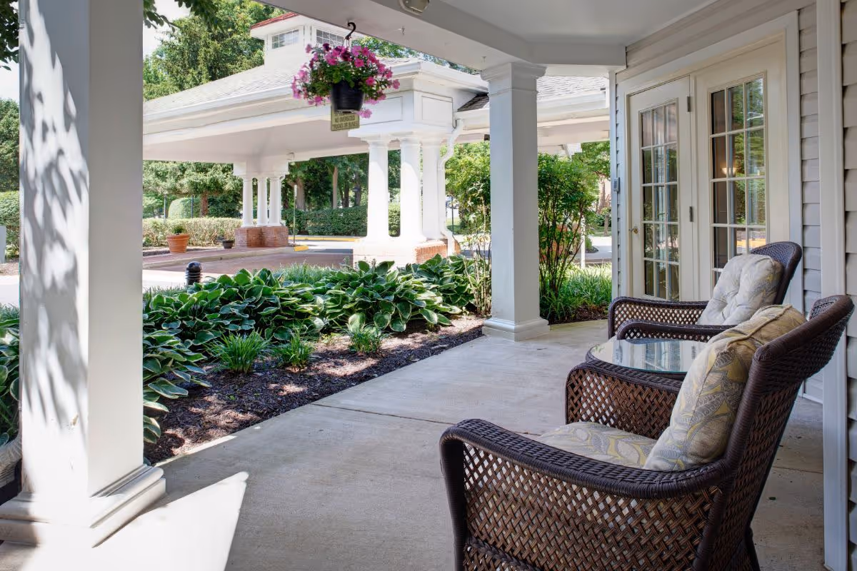 Covered outdoor patio area with two cushioned wicker chairs and a glass-top table, overlooking a garden bed with green plants and a white columned entrance structure in the background.