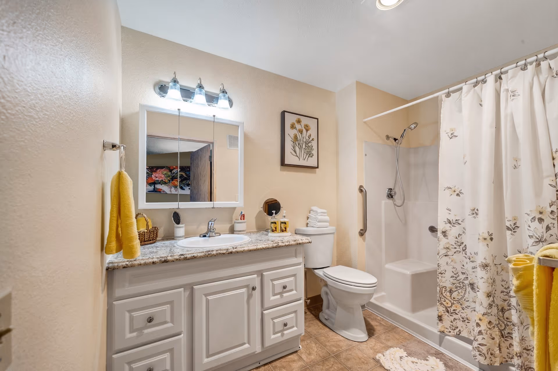 A clean and well-lit bathroom featuring a white vanity with a granite countertop and a sink, a mirror with three lights above it, a toilet with folded white towels on top, and a shower with a floral-patterned curtain. The walls are painted beige, and there is a framed floral artwork above the toilet. Yellow towels hang on a towel rack and a hook.