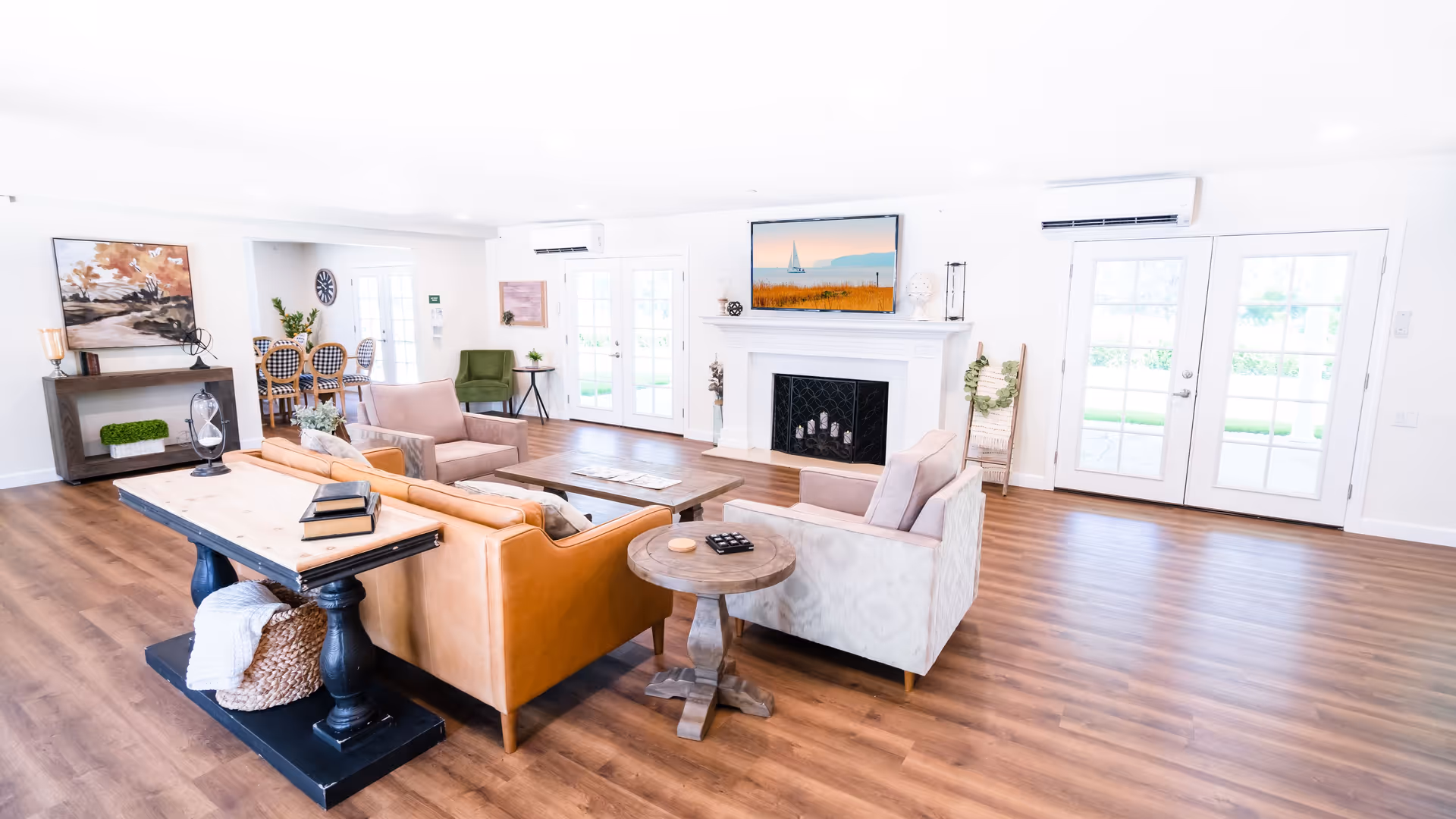 Bright and spacious senior living common area with wooden flooring, a tan leather sofa, two armchairs, a wooden coffee table, and a side table. The room features a white fireplace with a flat-screen TV mounted above it, large windows and glass doors letting in natural light, and a dining area with a table and chairs visible in the background.