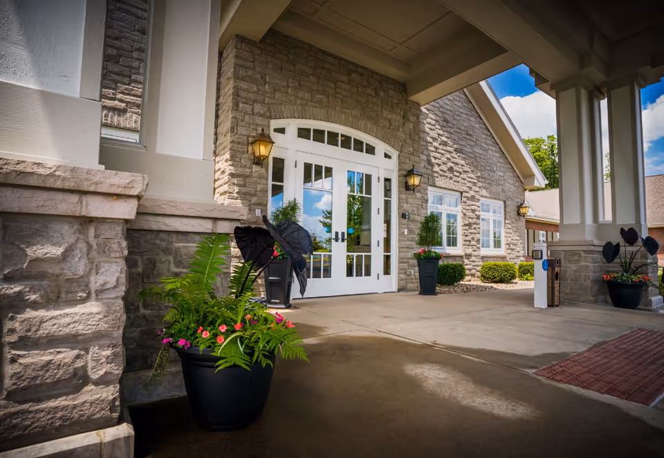 Entrance of The Inn at Ashland Woods showing a covered driveway with stone walls, white double doors with glass panels, outdoor wall lanterns, and large potted plants with green foliage and colorful flowers.