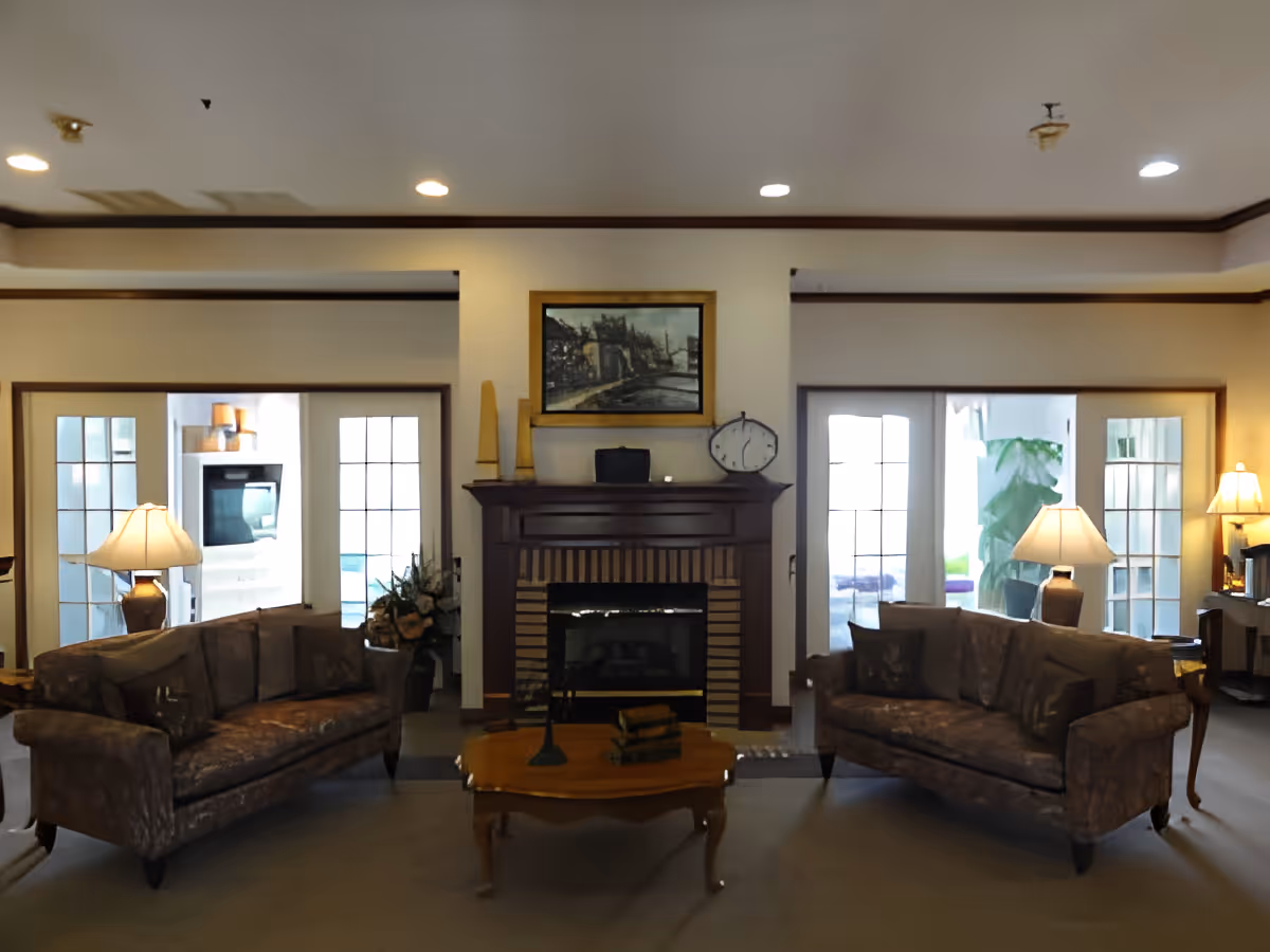 A cozy living room with two brown upholstered sofas facing each other, a wooden coffee table in the center with decorative items, and a fireplace with a mantel. The mantel holds a clock, two tall candles, and a framed painting above it. There are two table lamps on side tables next to each sofa, and French doors with glass panes lead to another room with visible plants and a fireplace.
