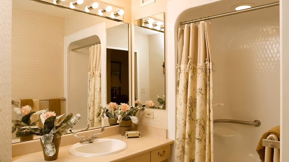 A bathroom with a beige countertop and sink, a large mirror with lights above it, a floral shower curtain partially covering a bathtub with a grab bar, and two potted plants with pink flowers on the counter. Towels are hanging on a rack on the wall.