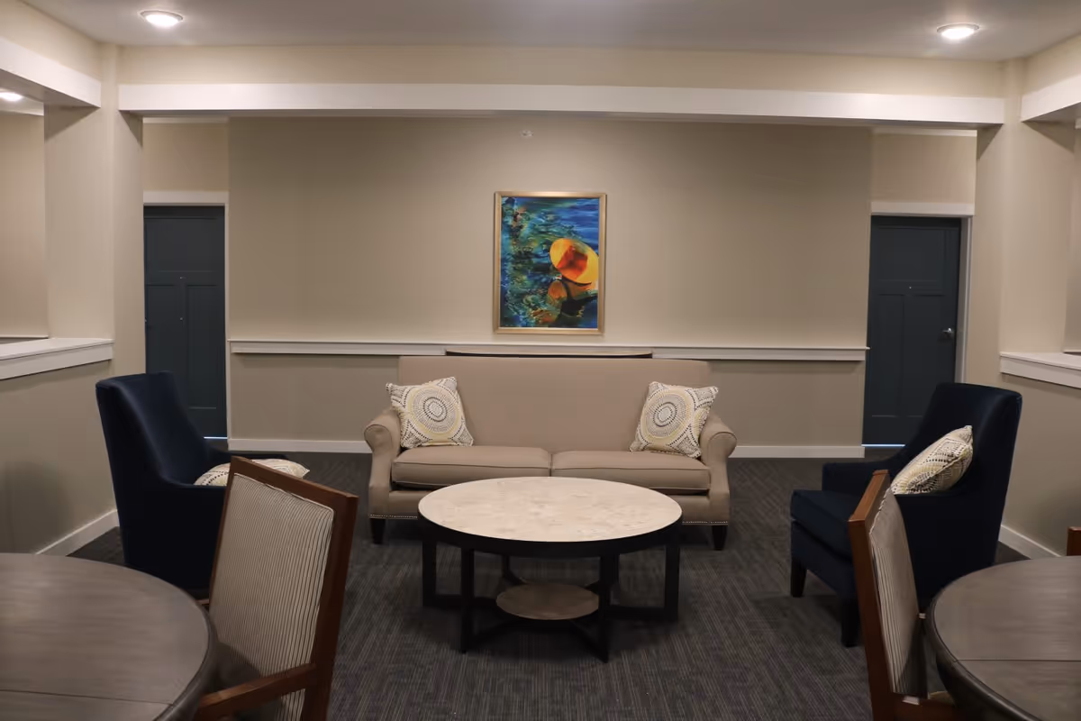 A cozy seating area in a senior living facility featuring a beige sofa with two patterned pillows, two dark blue armchairs each with a pillow, and a round coffee table in the center. The walls are painted light beige with a colorful framed painting of a person wearing a yellow hat above the sofa. There are two closed dark doors on either side of the sofa, and part of a round dining table with chairs is visible in the foreground.
