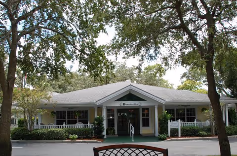 Front exterior view of Discovery Commons At Bradenton, a single-story building with a gray roof, white columns, and a covered entrance surrounded by trees and greenery.